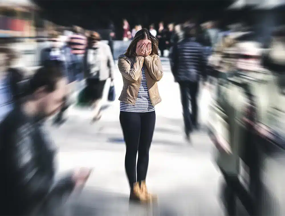 A person stands covering their face in a busy, blurred crowd outdoors during the day.