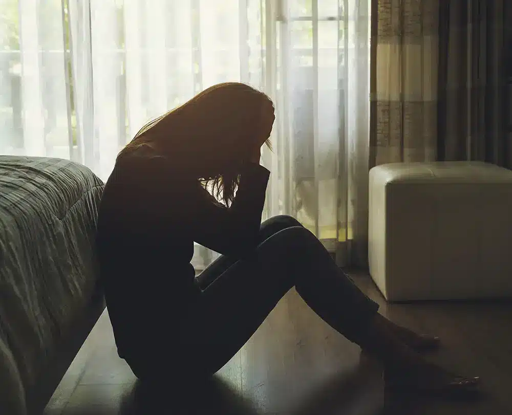 Person sitting on the floor by a bed, head down, in a dimly lit room with sheer curtains.